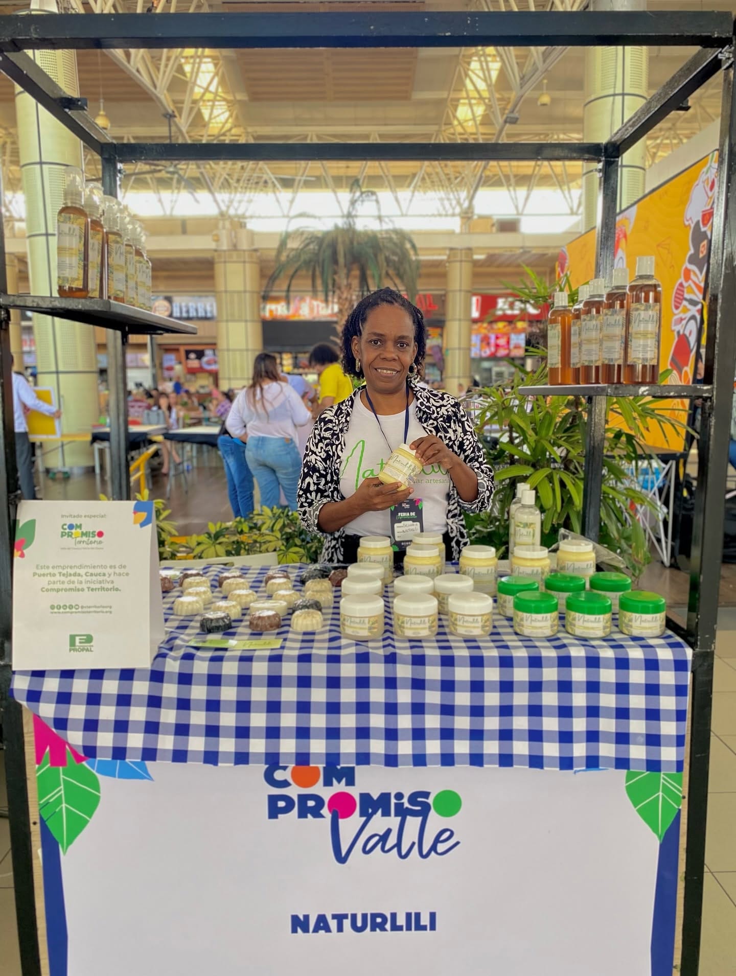 Liliana en su puesto de feria de Compromiso Valle, sosteniendo un producto Naturlili frente a la mesa de jabones y potes.